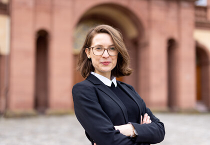 Svenja Behrendt trägt einen schwarzen Blazer, einen Pullover mit weißem Kragen und lächelt in die Kamera. Sie steht vor dem Schloss.