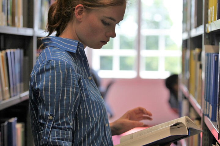 Studentin in der Bibliothek vor einem Regal, liest in Buch