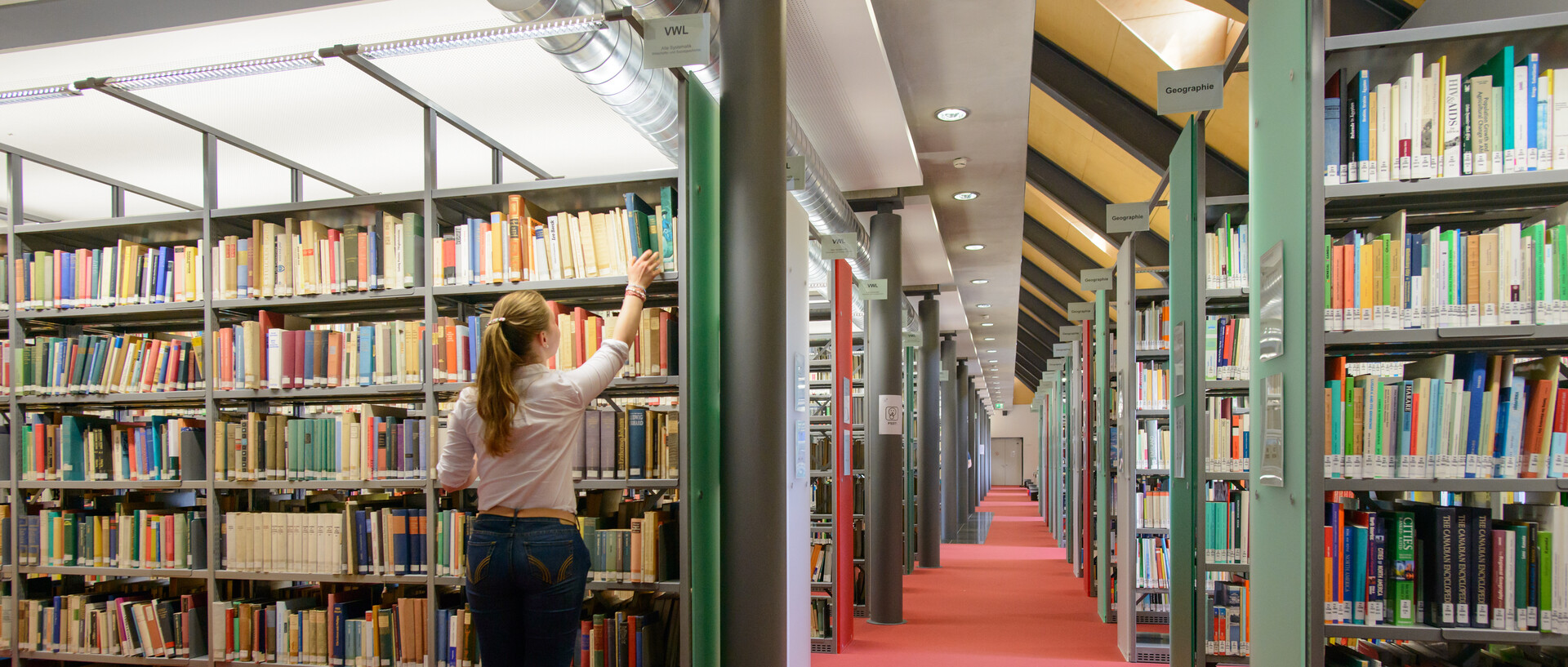 Bibliothek mit rotem Teppich und Bücherregalen, eine Studentin holt ein Buch aus einem Regal