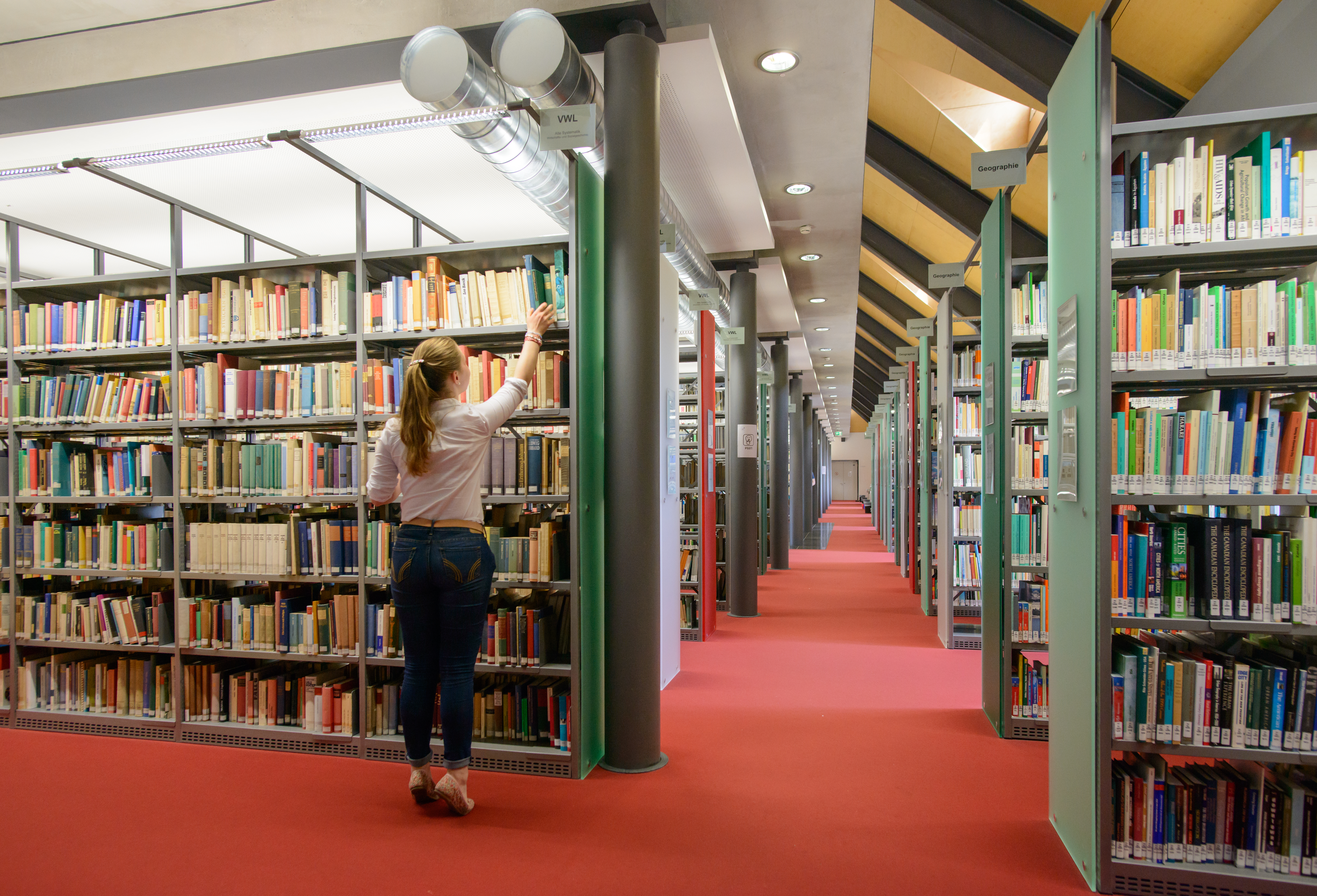 Bibliothek mit rotem Teppich und Bücherregalen, eine Studentin holt ein Buch aus einem Regal