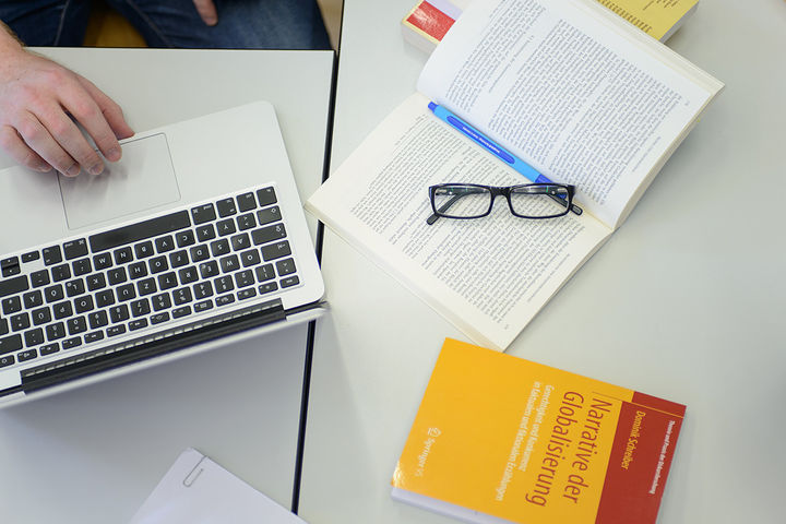 Auf einem Tisch liegen ein Laptop, ein Buch mit dem Titel "Narrative der Globalisierung" und ein aufgeschlagenes Buch mit Brille und Stift.