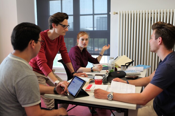 Studierende sitzen an einem Tisch mit Büchern und einem Laptop und diskutieren