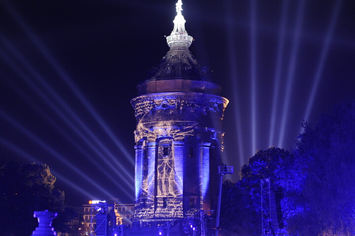 Der Mannheimer Wasserturm blau beleuchtet bei Nacht. Im Hintergrund mehrere blaue Scheinwerfer.