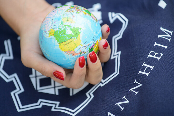 Eine Hand mit roten lackierten Fingernägeln hält einen kleinen Globus in der Hand. Dahinter ist das Logo der Universität zu sehen. 