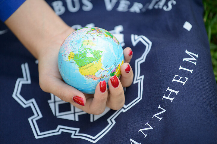 Hand mit rot lackierten Fingernägeln hält kleinen Globus in der Hand, dahinter ist das Logo der Universität zu sehen