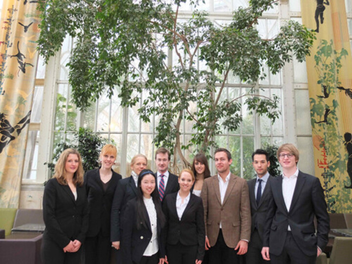 Gruppe von Studierender in formeller Kleidung vor  einem großen Fenster mit Pflanze
