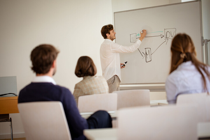 Forschende sitzen in einem Seminarraum an Tischen. Ein Forscher erklärt etwas an einem Whiteboard.