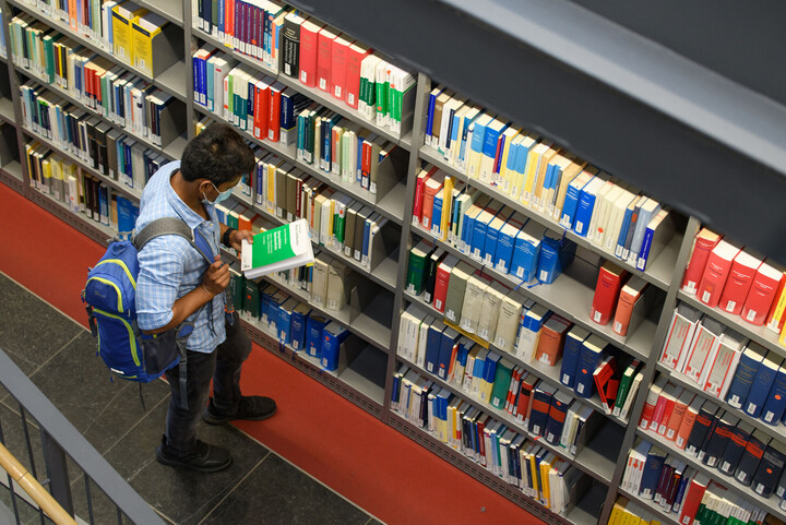 Ein Student mit Maske steht vor einem Bibliotheksregal und schaut sich ein Buchcover an.