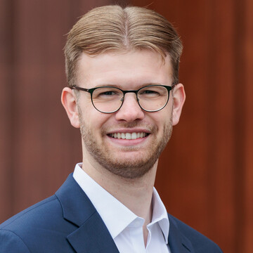 Portrait von Tim Gromes. Er trägt einen blauen Anzug mit weißem Hemd und steht vor einer brauen Tür der Universität.