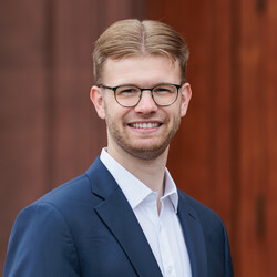 Portrait von Tim Gromes. Er trägt einen blauen Anzug mit weißem Hemd und steht vor einer brauen Tür der Universität.