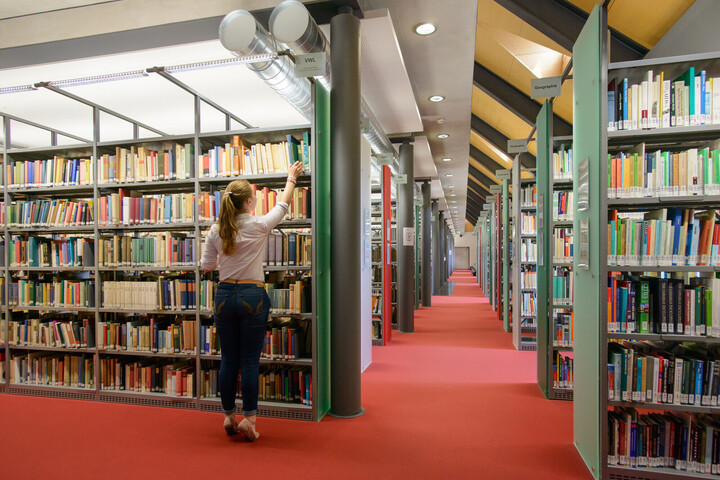 Eine Studentin stellt sich auf die Zehenspitzen, um aus dem obersten Bibliotheksregal ein Buch zu ziehen.