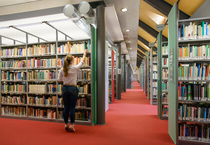 Eine Studentin stellt sich auf die Zehenspitzen, um aus dem obersten Bibliotheksregal ein Buch zu ziehen.