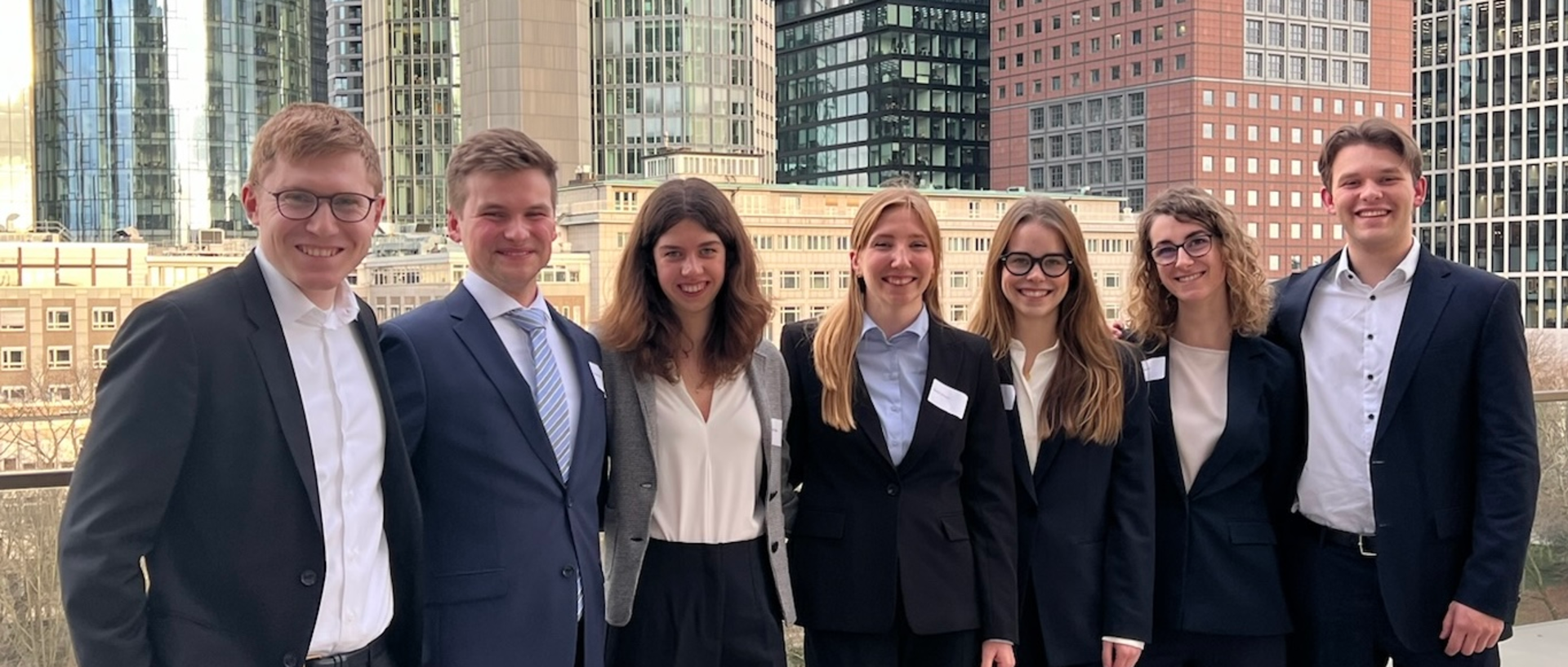 Eine Gruppe Studierender stehen in Anzügen auf einer Terrasse mit Blick auf die Frankfurter Skyline
