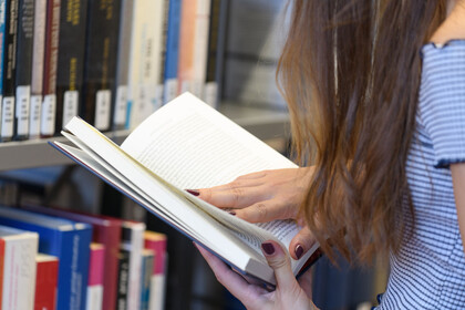 Nahaufnahme einer vor dem Buchregal stehenden Studentin, die in einem Buch liest.