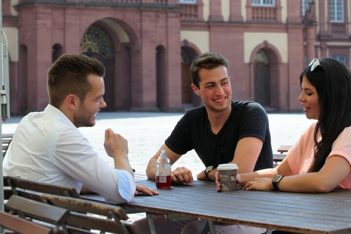 Drei Studierende sitzen an einem Tisch im Ehrenhof und unterhalten sich. 
