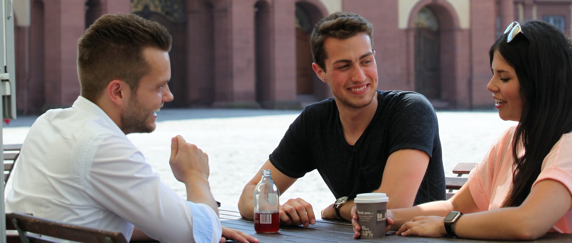 Drei Studierende sitzen an einem Tisch im Ehrenhof und unterhalten sich