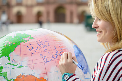 Lächelnde Frau in einem gestreiften Shirt auf dem Ehrenhof, schreibt mit einem Stift auf einen großen Globus "Hello World".