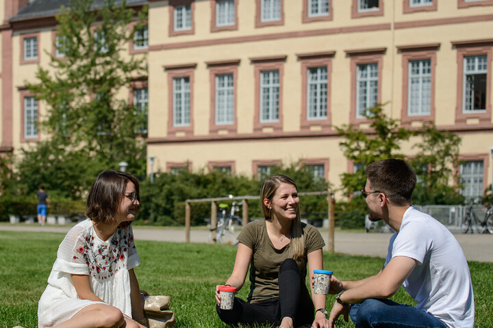 Zwei Studentinnen und ein Student sitzen auf einer grünen Wiese. Sie tragen frühlingshafte Kleidung und halten Kaffee-To-Go-Becher in ihren Händen.