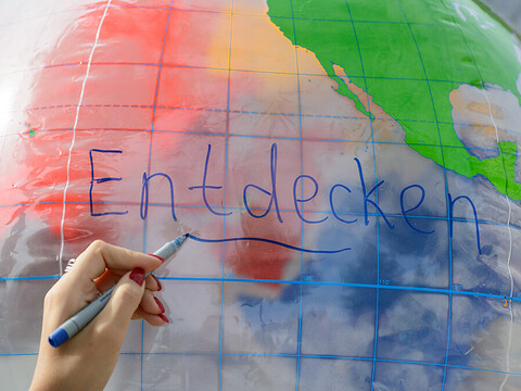 Eine Hand schreibt das Wort "entdecken" auf einen aufgeblasenen Globus.