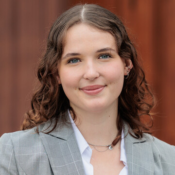 Portrait von Sara Bonhoff. Sie trägt einen hellgrauen Anzug mit weißem Hemd und steht vor einer brauen Tür der Universität.