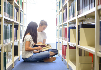 Zwei Studentinnen sitzen in der Bibliothek auf dem Boden vor dem Regel und blättern in Büchern