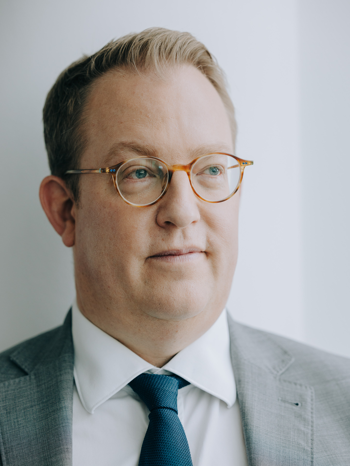 Porträtaufnahme. Er trägt eine Brille und Krawatte. Er schaut nach rechts. 