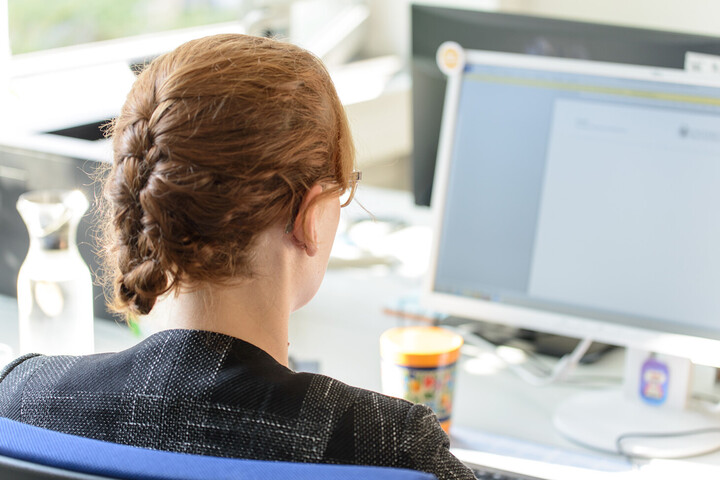 Eine Frau sitzt ist von hinten zu sehen, wie sie an einem Computer arbeitet