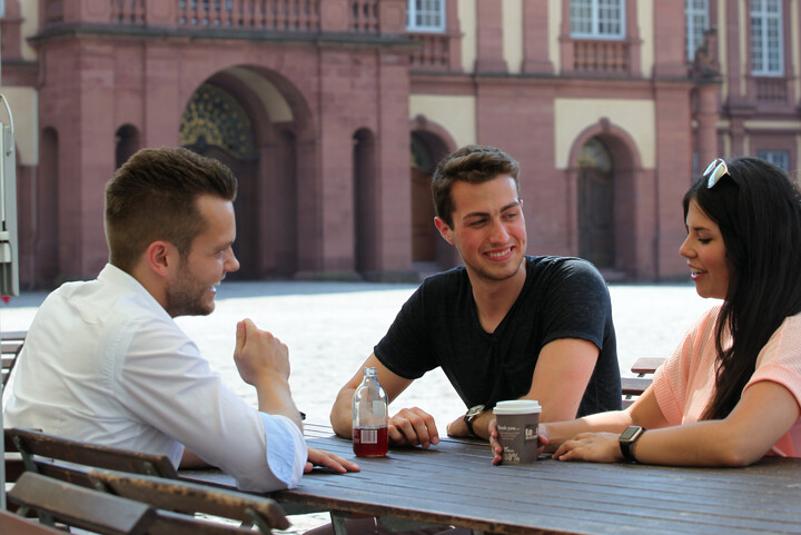 Drei Studierende sitzen an einem Tisch in der Sonne und unterhalten sich