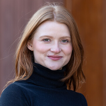 Portrait von Astrid Megner. Sie trägt einen schwarzen Rollkragenpullover und steht vor einer brauen Tür der Universität.