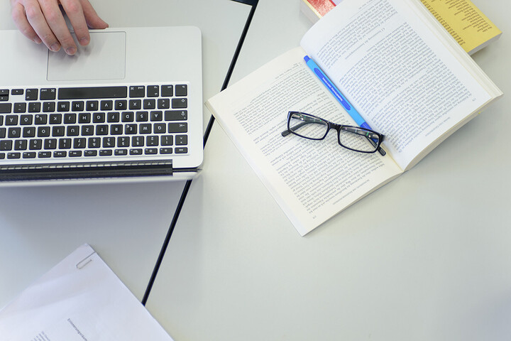 Ein Laptop, ein aufgeschlagenes Buch und eine Brille liegen auf dem Tisch.