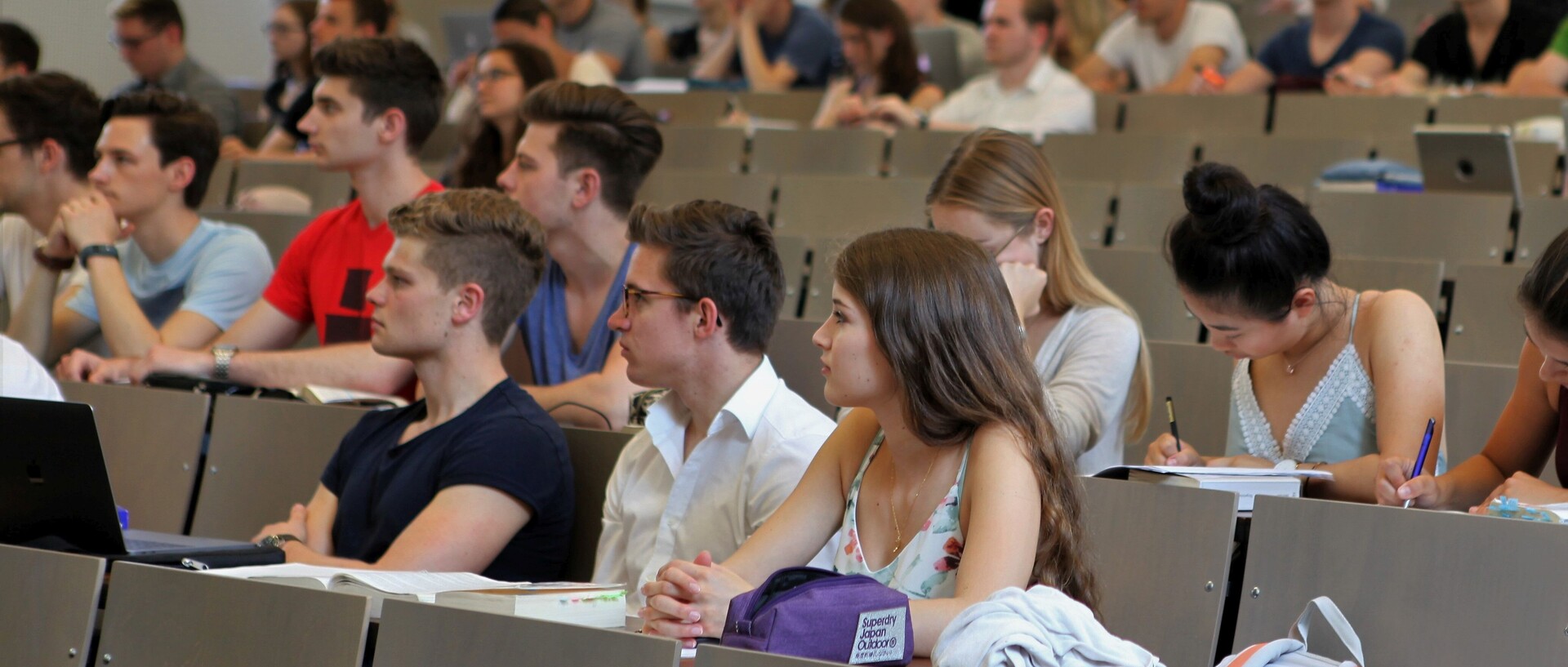 Studierende sitzen in einem Hörsaal. Die Studierenden tragen kurzärmlige Oberteile und vor ihnen liegen Laptops, Tablets oder Notizblöcke.
