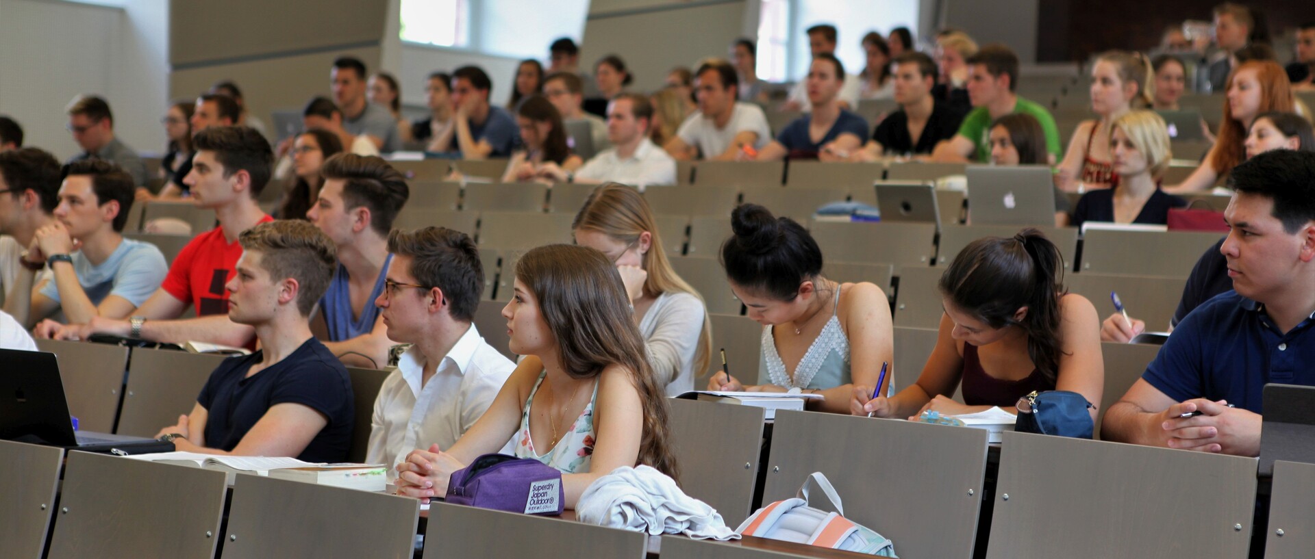 Studierende sitzen in einem Hörsaal. Die Studierenden tragen kurzärmlige Oberteile und vor ihnen liegen Laptops, Tablets oder Notizblöcke.