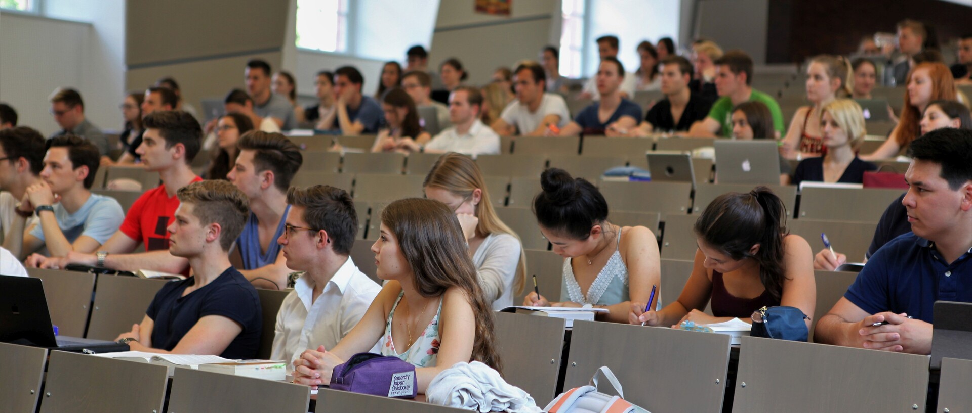 Studierende sitzen in einem Hörsaal. Die Studierenden tragen kurzärmlige Oberteile und vor ihnen liegen Laptops, Tablets oder Notizblöcke.