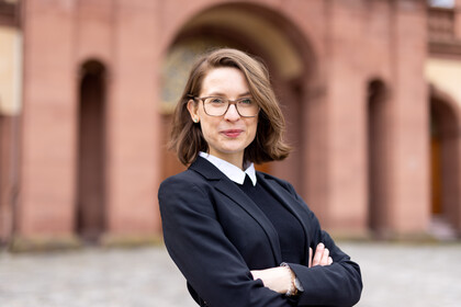 Svenja Behrendt trägt einen schwarzen Blazer, einen Pullover mit weißem Kragen und lächelt in die Kamera. Sie steht vor dem Schloss.