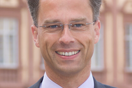 Portrait von Georg Bitter. Er tträgt ein weißes Hemd, eine lila-farbene Krawatte sowie einen grauen Blazer und steht vor dem Schloss.