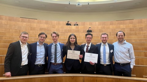 Eine Gruppe Studierender in Anzügen vor einem Saal mit leeren Stühlen. Zwei Personen halten ein Dokument in den Händen.