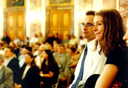 Eine Frau und ein Mann stehend im Vordergrund. Mehrere Menschen sitzend im Hintergrund.