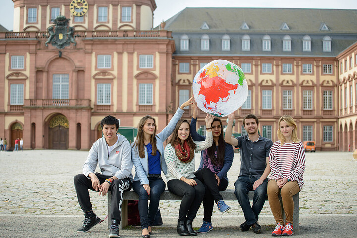 Gruppe Studierender sitzt auf einer Bank vor dem Schloss und halten einen aufgeblasenen Globus in die Höhe