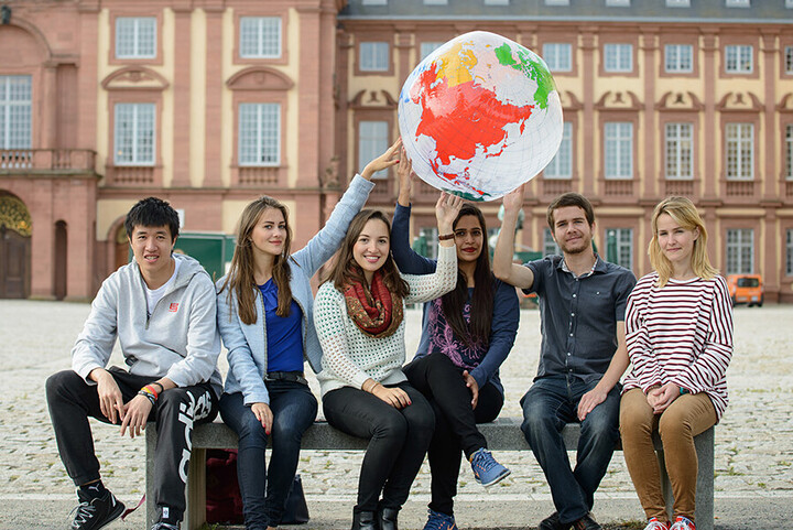 Studierende sitzen auf einer Bank vor dem Schloss und halten einen aufgeblasenen Globus in die Luft.