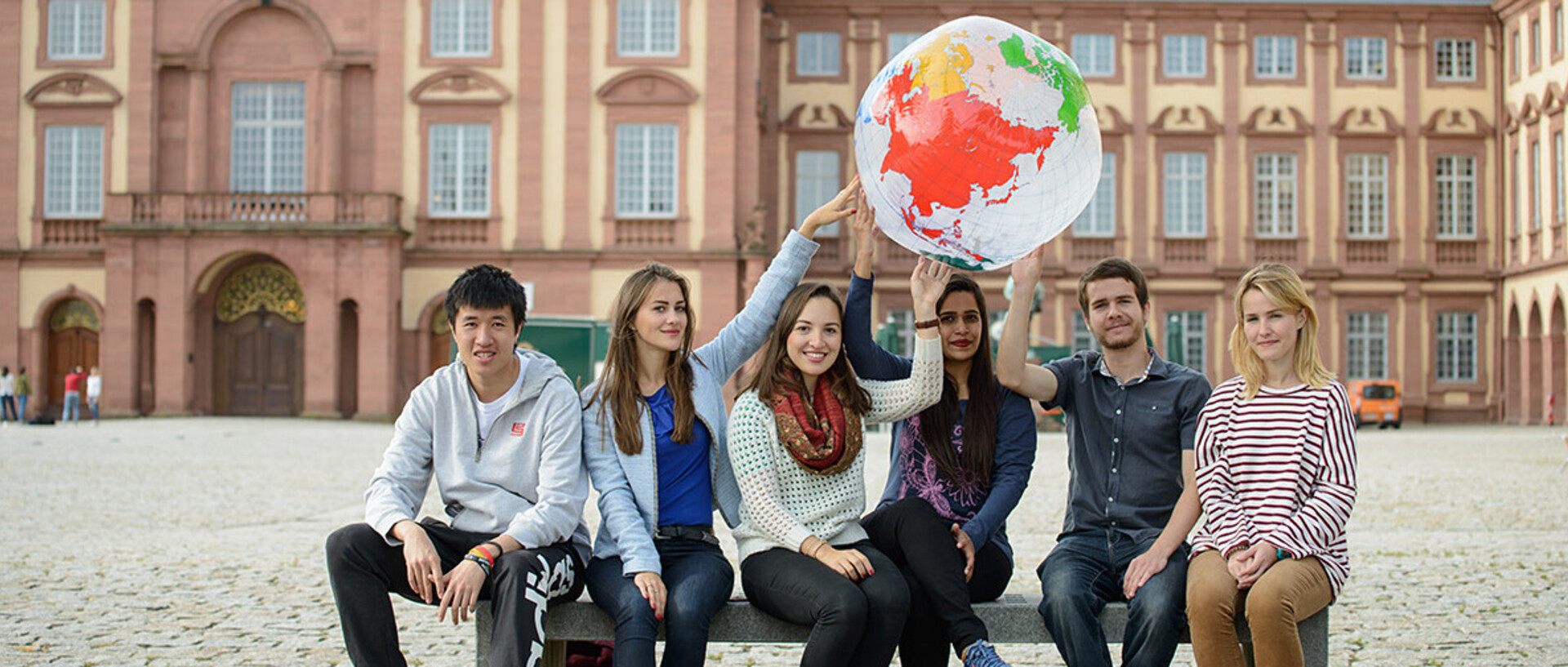 Eine Gruppe Studierender sitz auf einer Bank vor dem Schloss und hält einen aufgeblasenen Globus in die Luft