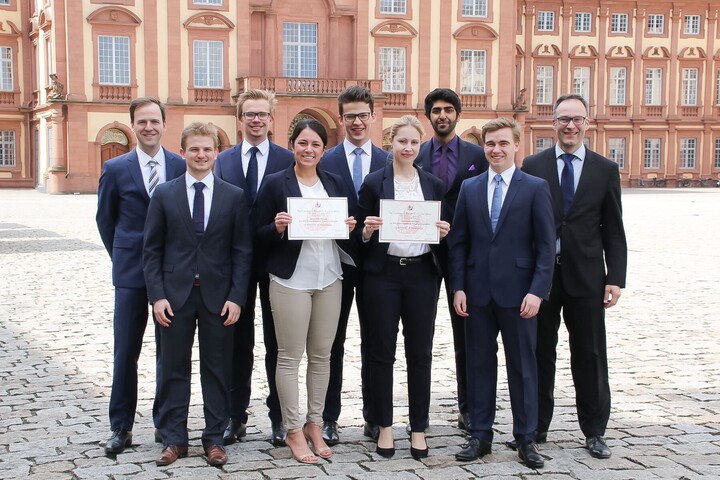 Eine Gruppe Studierender in Anzügen steht vor dem Schloss und hält gemeinsam ein Dokument