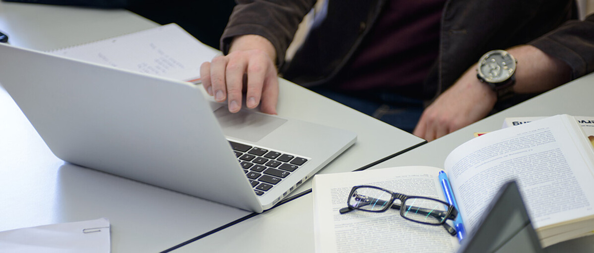 Ein offener Laptop mit einer Hand, die an der Tastatur arbeitet. Daneben ein aufgeschlagenes Buch mit Brille darauf