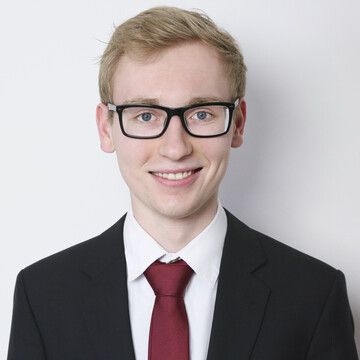 Lennard Streb hat kurze blonde Haare. Er trägt eine schwarze eckige Brille, ein weißes Hemd, eine rote Krawatte und ein schwarzes Jackett.