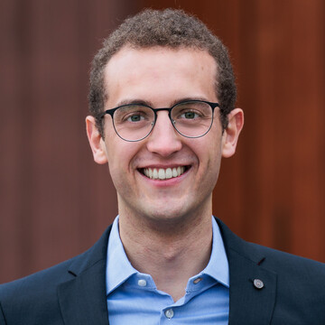 Portrait von Lukas Romberg. Er trägt einen Anzug mit blauem Hemd und steht vor einer brauen Tür der Universität.