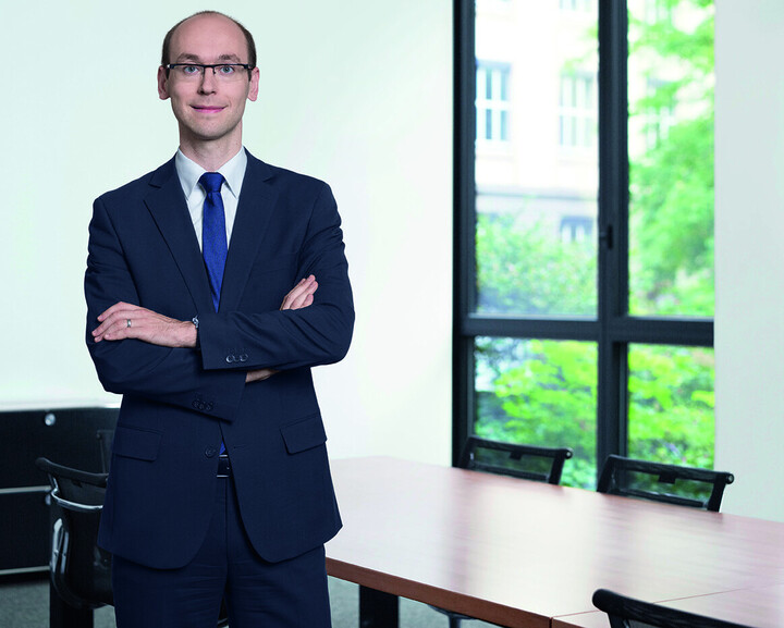 Simon Apel standing in black suit and tie in front of a window