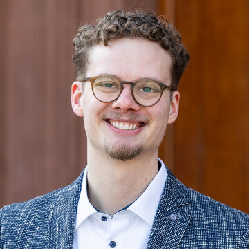 Portrait von Tim Vollmer. Er trägt einen Anzug mit weißem Hemd und steht vor einer brauen Tür der Universität.