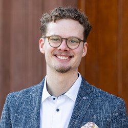 Portrait von Tim Vollmer. Er trägt einen Anzug mit weißem Hemd und steht vor einer brauen Tür der Universität.