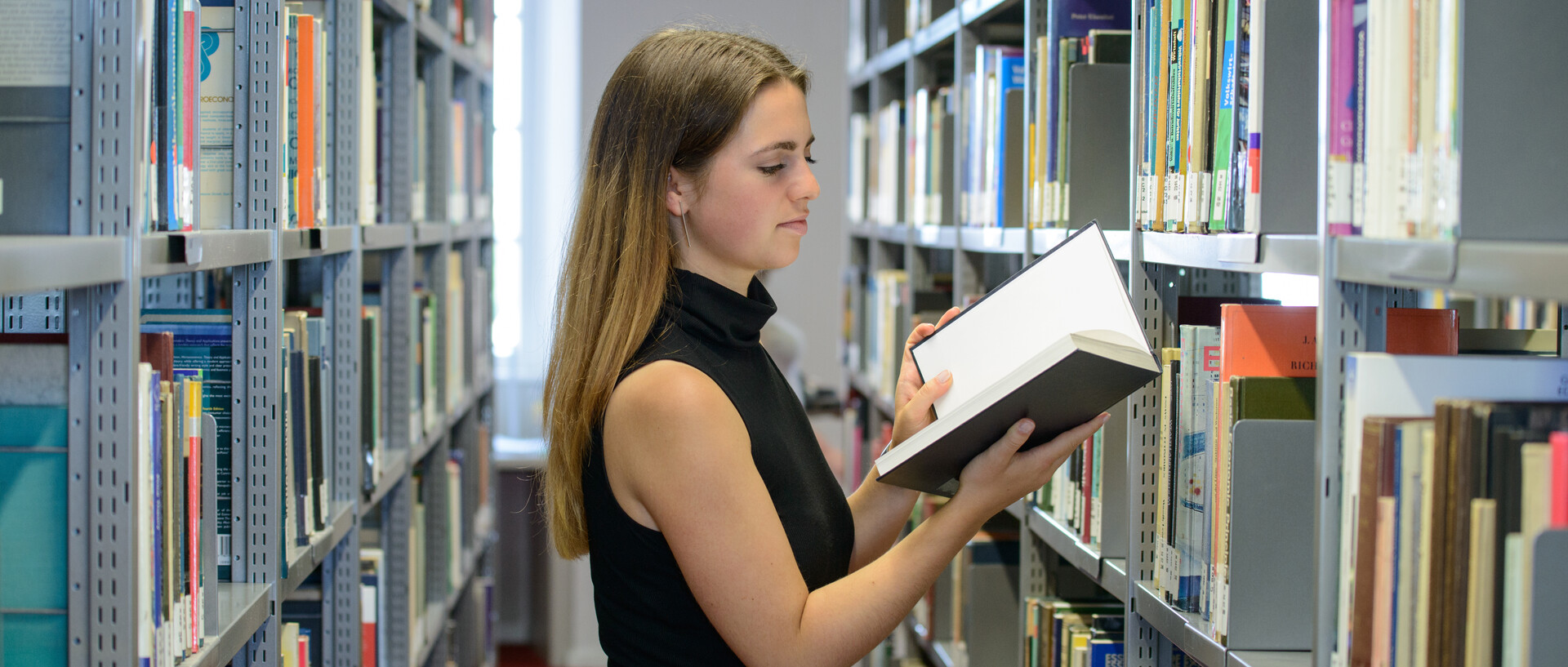 Dekoratives Bild, auf welchem eine Frau ein Buch zwischen den Regalen der Bibliothek liest.