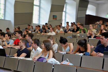 Studierende sitzen in einem Hörsaal. Die Studierenden tragen kurzärmlige Oberteile und vor ihnen liegen Laptops, Tablets oder Notizblöcke.