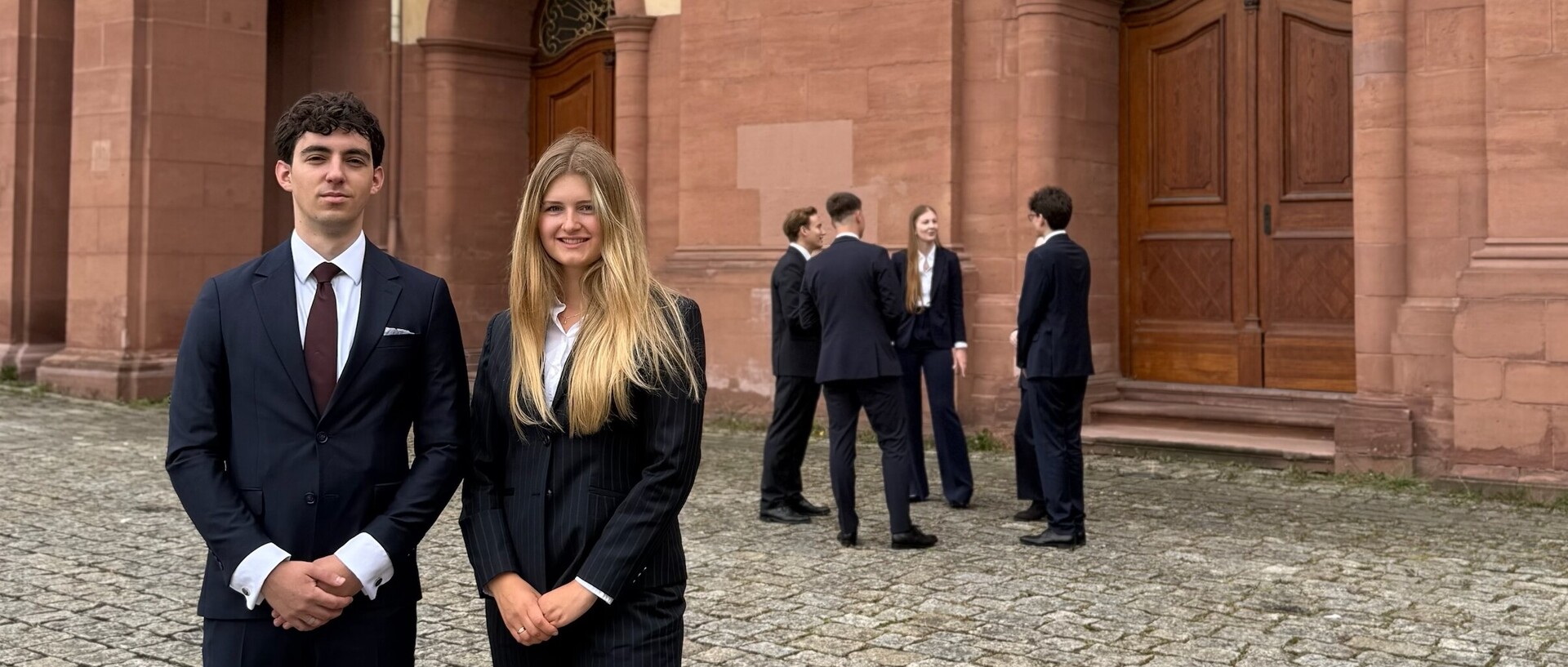 Zwei Studierende stehen lächelnd im Anzug im Vordergrund, während eine weitere Gruppe Studierender sich im Hintergrund unterhält.